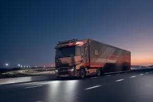 A Renault Trucks T High tractor unit pulling a motor racing semi-trailer is travelling along a motorway at dusk, near a floodlit circuit.
