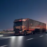 A Renault Trucks T High tractor unit pulling a motor racing semi-trailer is travelling along a motorway at dusk, near a floodlit circuit.