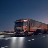 A Renault Trucks T High tractor unit pulling a motor racing semi-trailer is travelling along a motorway at dusk, near a floodlit circuit.
