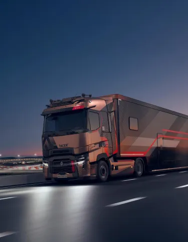 A Renault Trucks T High tractor unit pulling a motor racing semi-trailer is travelling along a motorway at dusk, near a floodlit circuit.